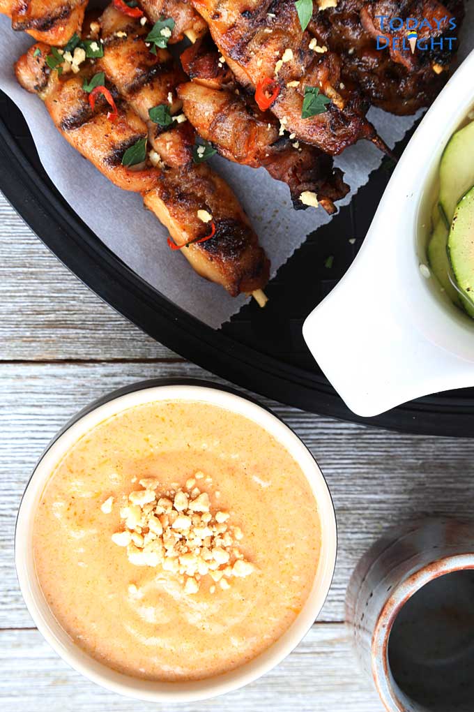 Chicken satay on a platter with lime wedges, peanut sauce in a bowl and a cucumber salad in a bowl.