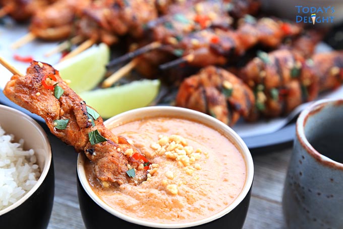 Chicken satay dipped in peanut sauce and in the background is a platter of chicken satay with lime wedges and rice ina bowl.