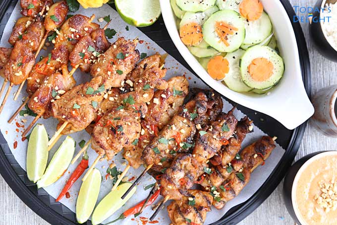 A platter of chicken satay with lime wedges and kaffir fruit, peanut sauce in a bowl, cucumber salad and rice.
