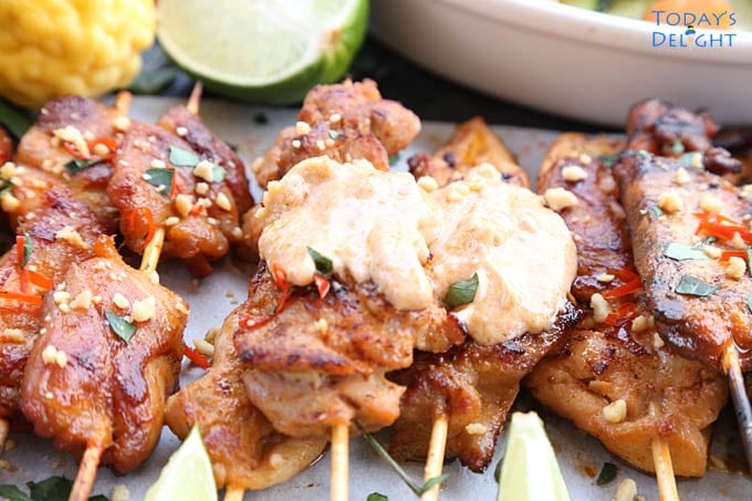 A platter of chicken satay with two skewers with peanut sauce. Lime wedges and kaffir fruit around the platter and cucumber salad on the side.