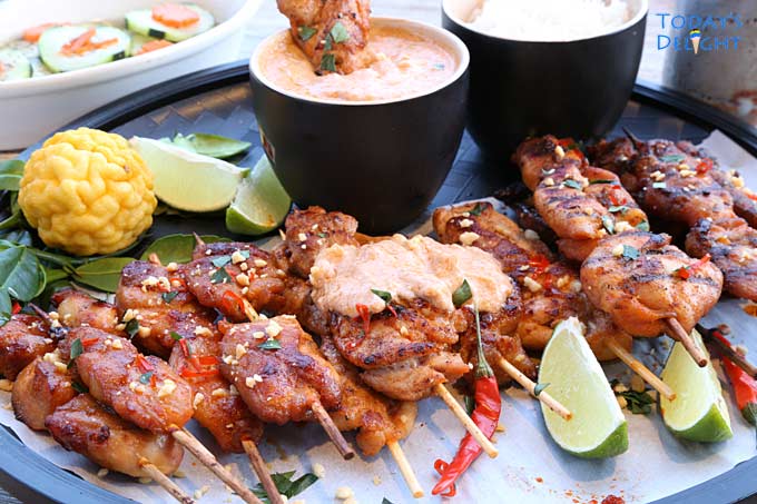 A platter of chicken satay with lime wedges and kaffir fruit, peanut sauce in a bowl, cucumber salad and rice.