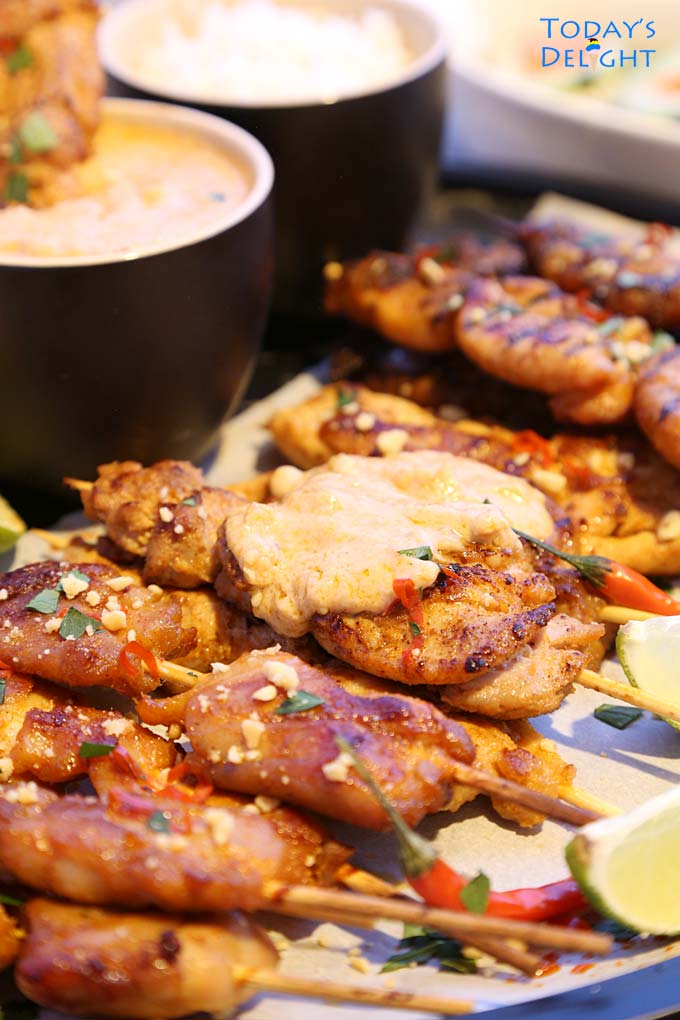 Chicken satay skewer dipped in peanut sauce and a platter of chicken satay with two skewers with peanut sauce. Lime wedges and kaffir fruit around the platter and cucumber salad on the side.