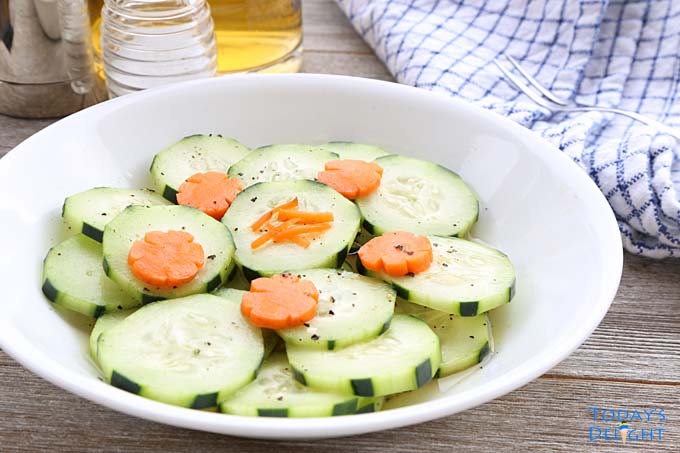 Sliced cucumber in a sweet sauce topped with decorative carrots. An appetizer fork, salt and napkin on the table.