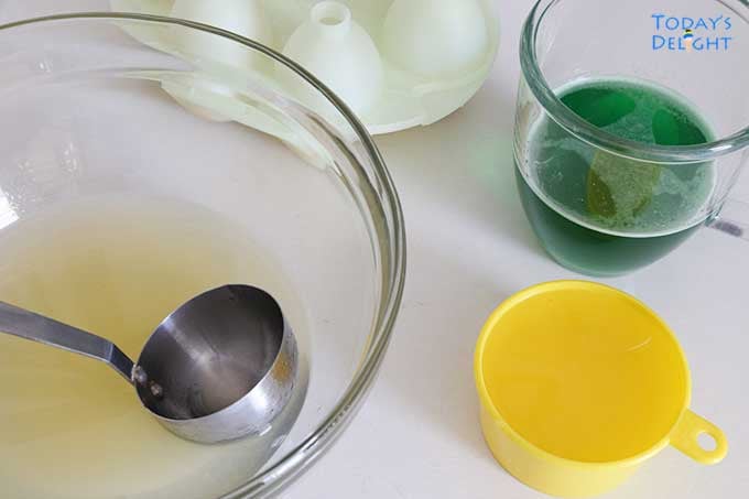 Green gelatin mixture in a cup, fruit cocktail juice in a bowl, 6 egg molds and a yellow measuring cup.