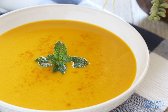 Kabocha soup garnished with mint leaves and a pinch of cayenne pepper. A vase in the background.