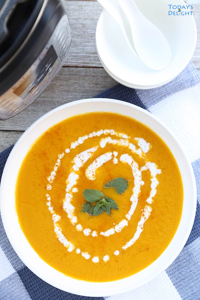 Kabocha squash soup garnished with mint leaves at the center and a swirl of coconut milk around the mints leaves. On the background is an instant pot and 2 bowls with spoons.
