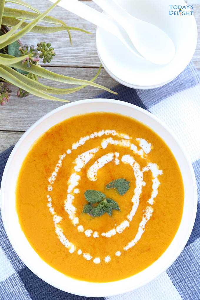 Kabocha squash soup garnished with mint leaves at the center, pinch of cayenne pepper and a swirl of coconut milk around the mints leaves. In the background, a vase and 2 bowls with spoons.