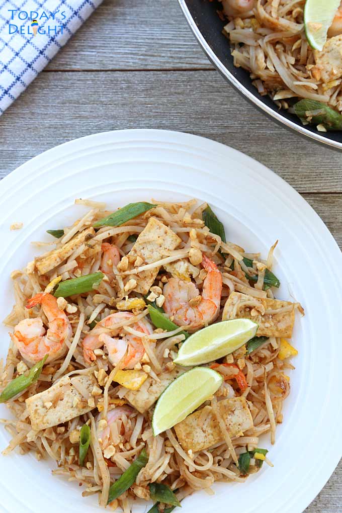 Shrimp pad Thai with tofu, bean sprouts, green onions, chopped peanuts and wedges of lime. Pad Thai skillet and napkin in the background.