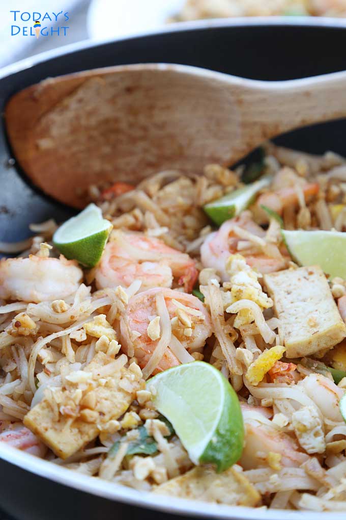 Pad Thai noodles with shrimp, tofu, bean sprouts, lime wedges, and green onions in a skillet with big wooden serving spoon.