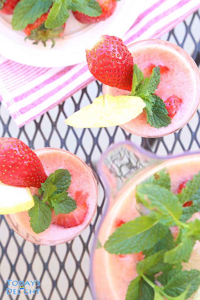 A pitcher and two glasses of Strawberry Lemonade Drink garnished with slice of strawberry, lemon and mint leaves.