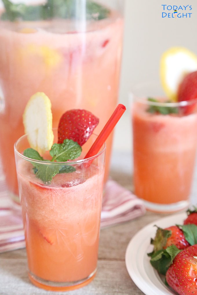Two glasses of homemade Strawberry Lemonade Drink garnished with strawberry, lemon and mint leaves and a pitcher and some whole strawberries on the side.