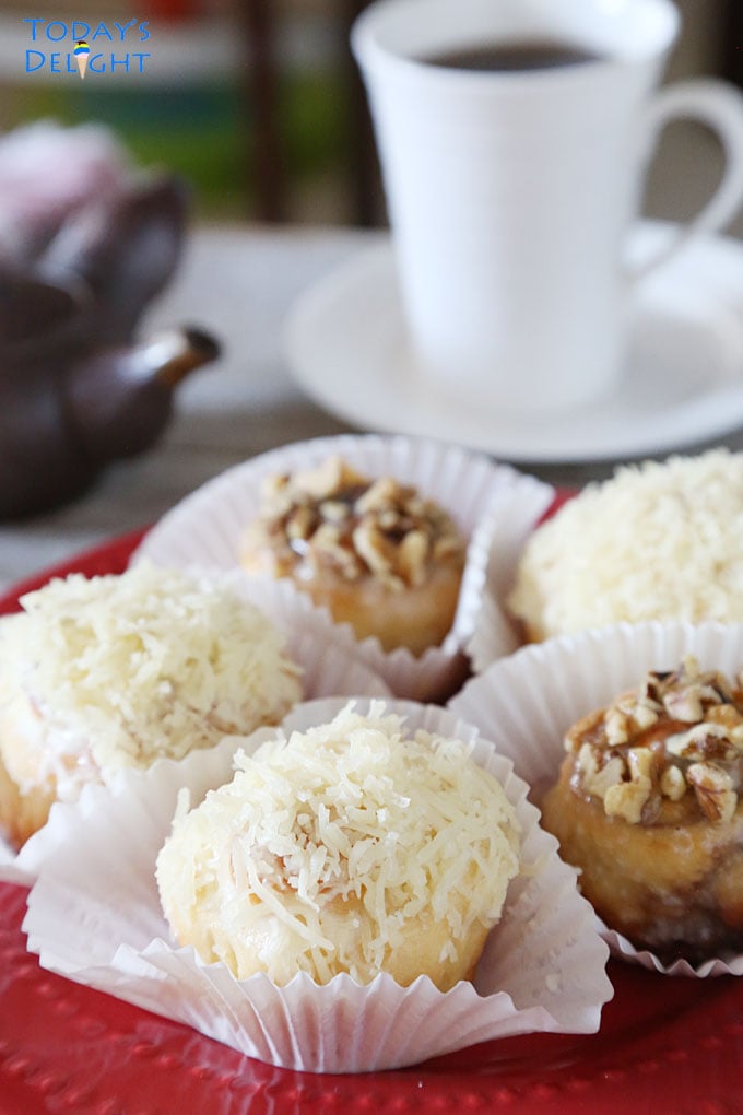 Three ensaymada sweet rolls topped with butter, sugar, and grated cheese and two cinnamon topped with nuts on cupcake liners are on a plate. A cup of coffee and creamer on the side.