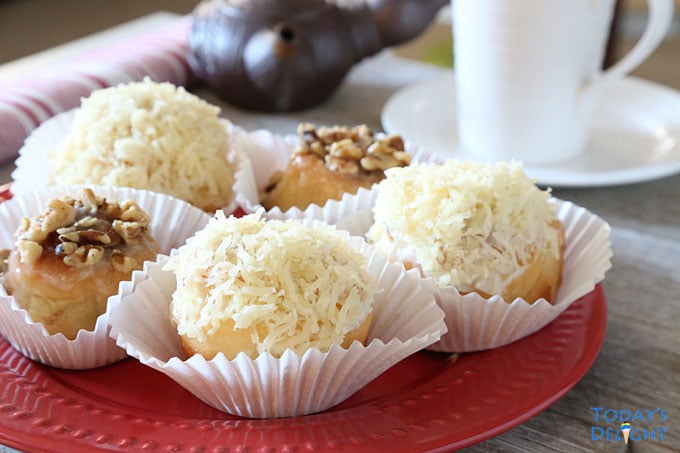 Three ensaymada sweet rolls topped with butter, sugar, and grated cheese and two cinnamon topped with nuts on cupcake liners are on a plate. A cup of coffee and creamer on the side.