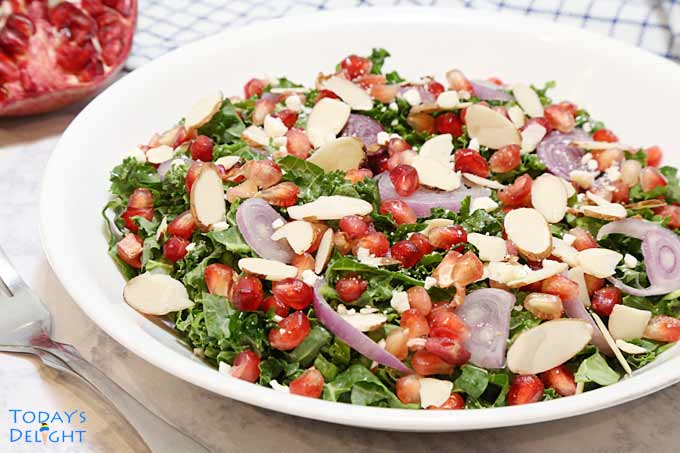 Pomegranate kale salad with shallots, shaved almonds, and feta cheese on a plate, pomegranate fruit on the side, and a fork.