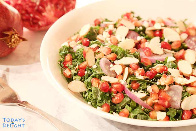 Pomegranate kale salad with shallots, shaved almonds, and feta cheese on a plate, pomegranate fruit on the side, and a fork.