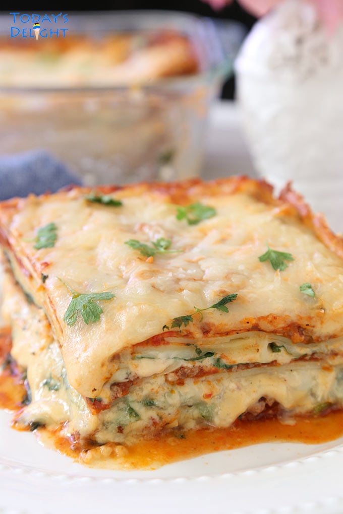 A slice of baked lasagna garnished with fresh parsley leaves on a plate, a vase and the lasagna casserole pan in the background.