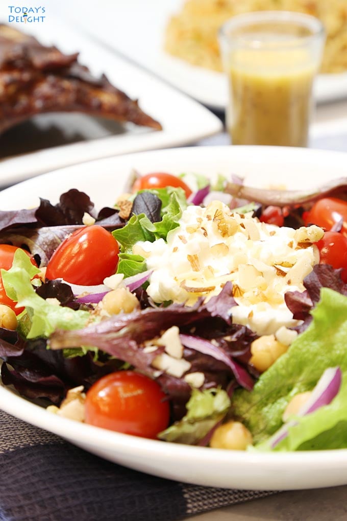 Salad made of mixed greens, chickpeas, red onion, cherry tomatoes, nuts and dressing on a plate.