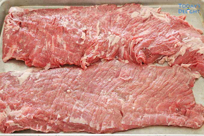 2 big slices of uncooked flank steak on a tray.