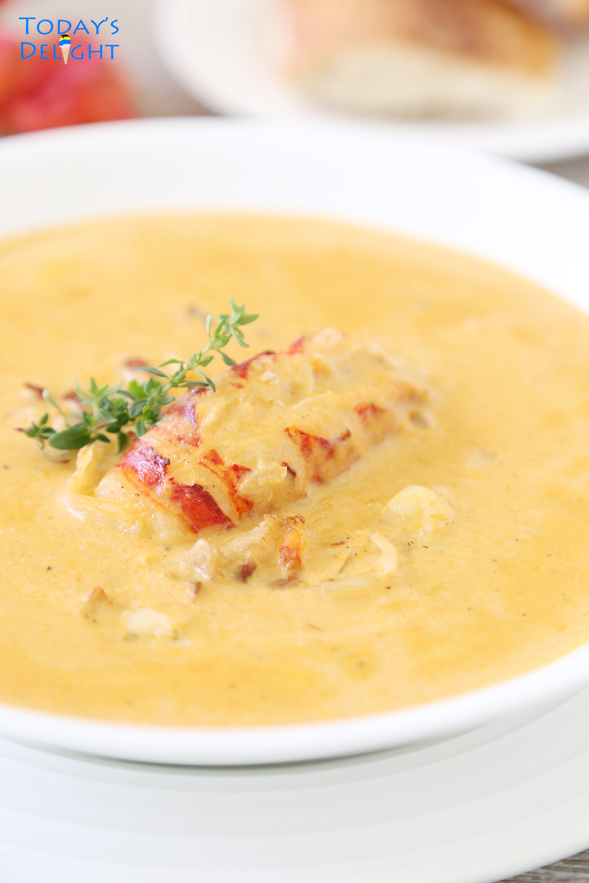 Lobster Bisque soup in a shallow bowl garnished with chunks and whole lobster tail and fresh thyme and slices of French bread.