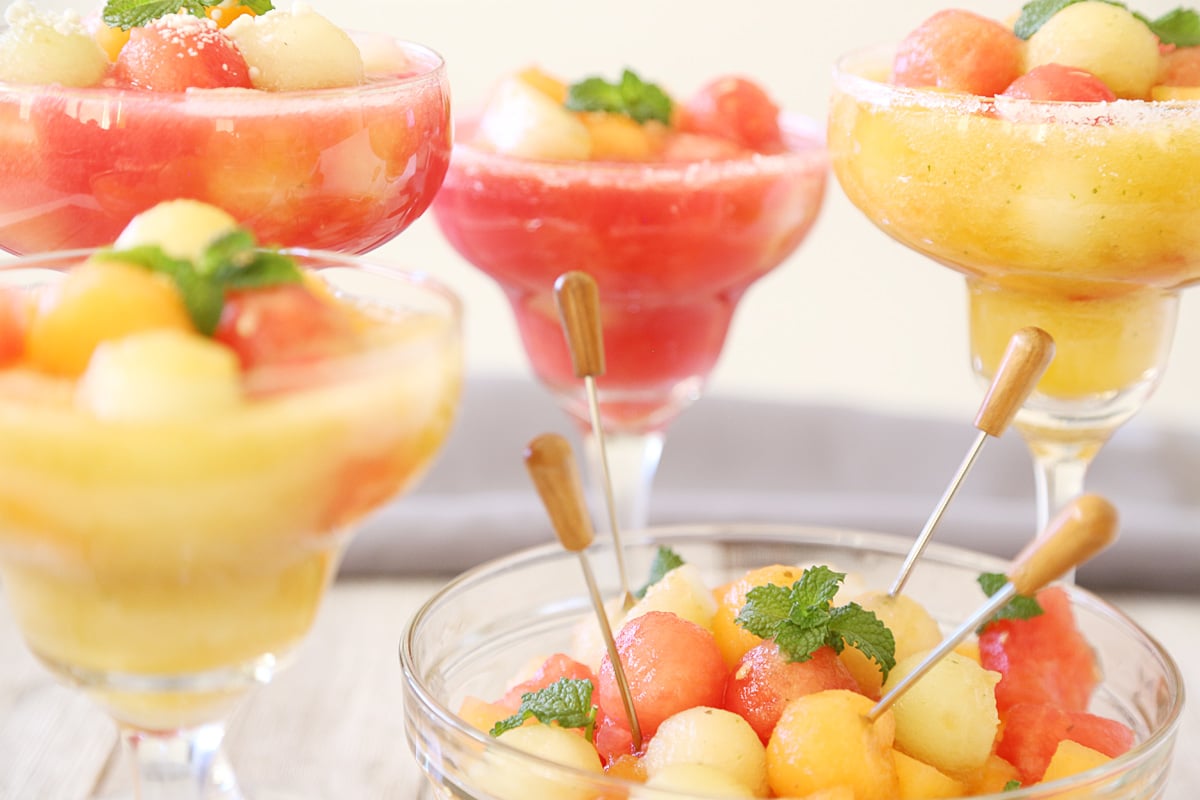 A big bowl of melon balls with cocktail sticks and 4 glasses of melon ball drink with mint.