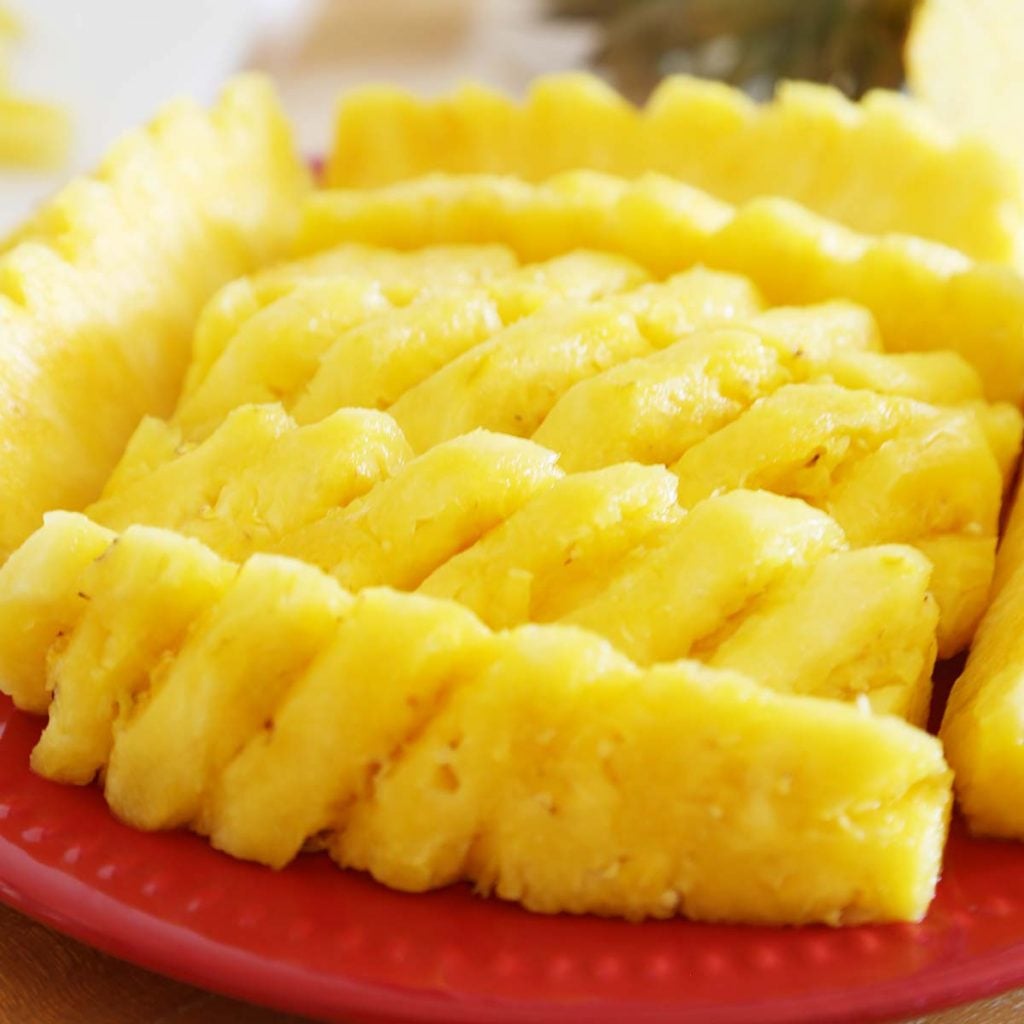 Fresh pineapple peeled, eyes removed decoratively, and sliced vertically into pieces on a red plate.