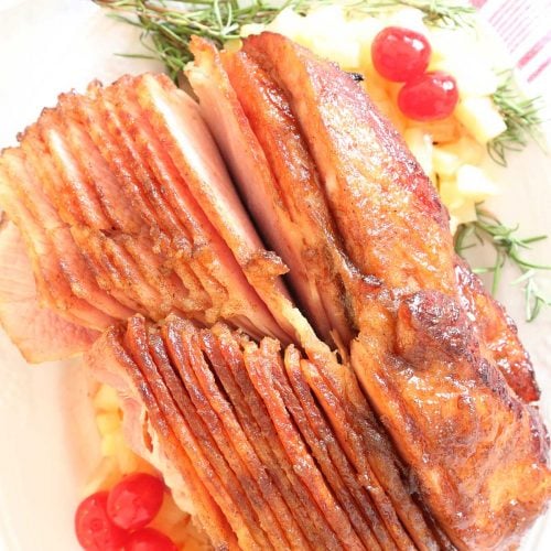 Pineapple honey glazed ham on a platter with rosemary, pineapple tidbits, cherries and side of sauce.