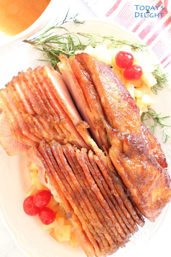 Pineapple honey glazed ham on a platter with rosemary, pineapple tidbits, cherries and side of sauce.
