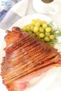 Glazed honey baked ham in a platter with green grapes and ham sauce on the side.