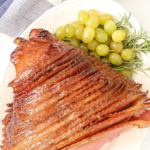 Glazed honey baked ham in a platter with green grapes and ham sauce on the side.