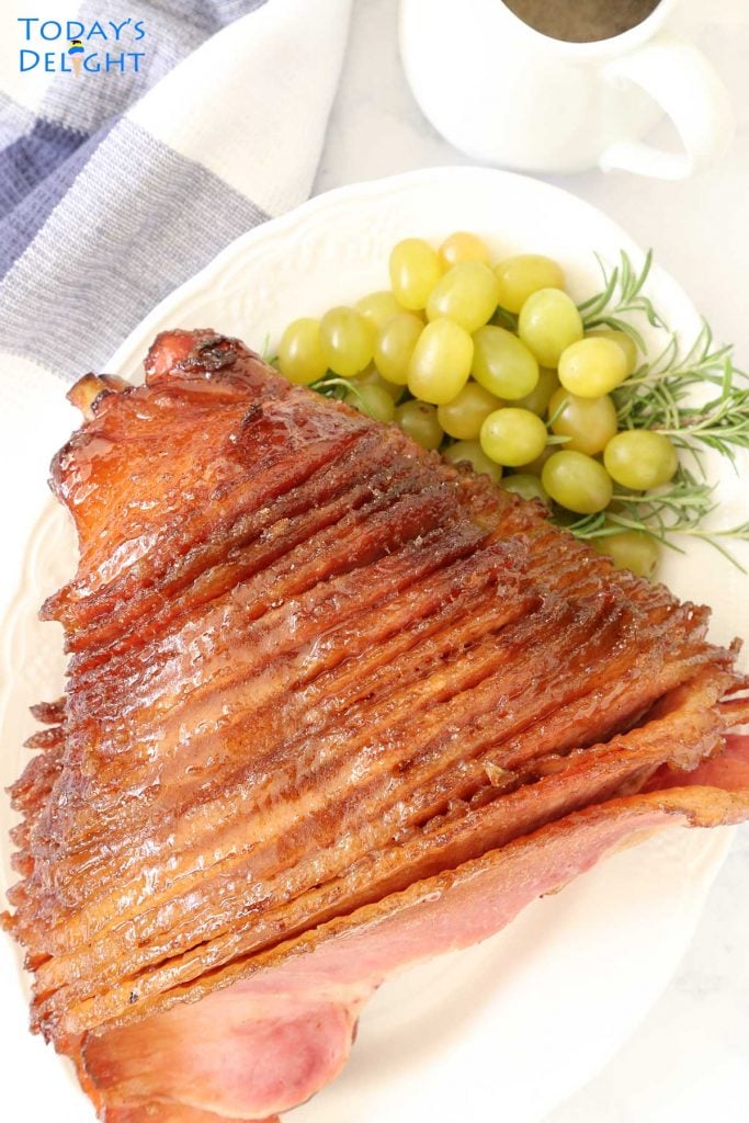 Glazed honey baked ham in a platter with green grapes and ham sauce on the side.