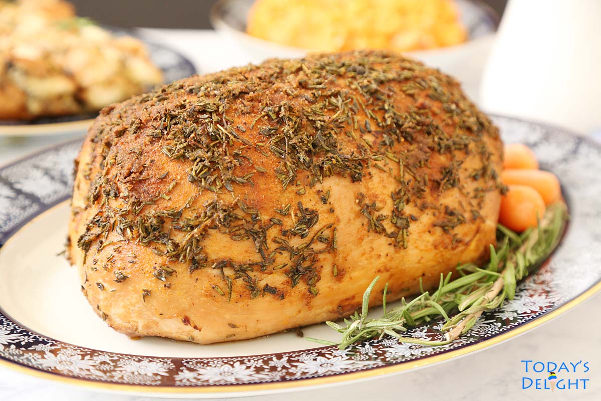 Seasoned fully cooked boneless turkey breast in a platter with fresh rosemary and baby carrots, turkey gravy and side dishes.