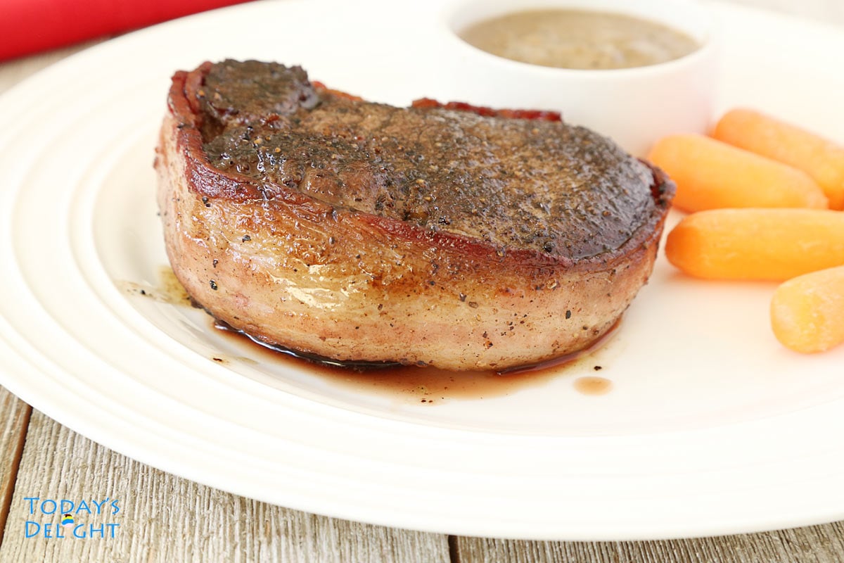 Filet mignon steak wrapped in bacon with a side of mushroom sauce and baby carrots.