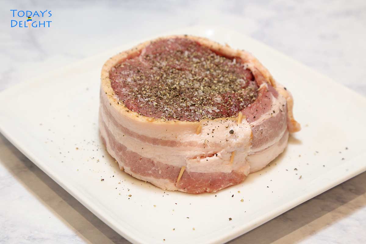 Uncooked bacon wrapped filet mignon seasoned with salt and pepper.