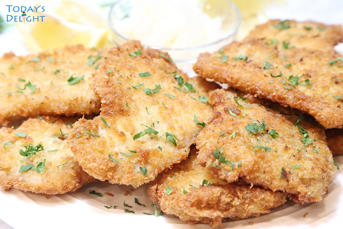 Crispy fried breaded pork cutlets garnished with chopped parsley on a platter with lemon wedges and sauerkraut on the side.
