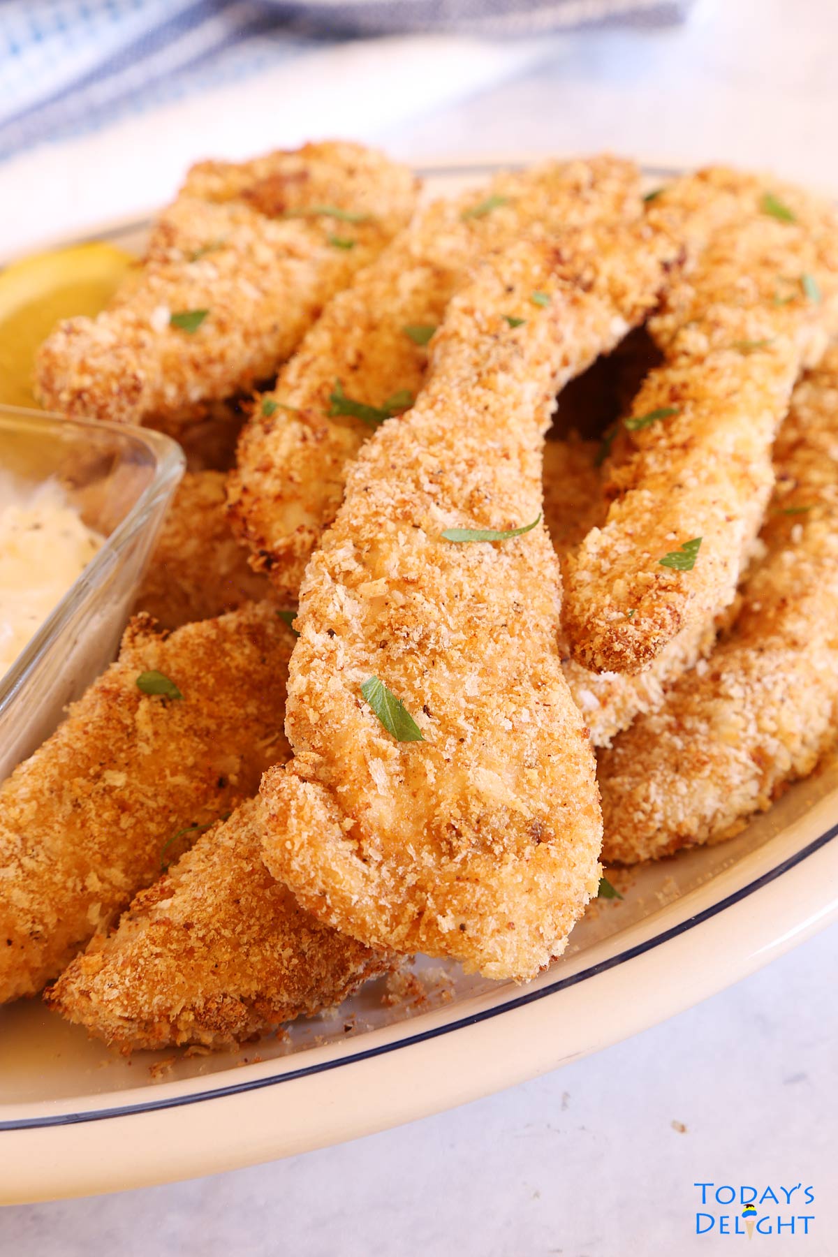 Crispy chicken tenders in a platter garnished with fresh parsley flakes and a side of sauce and lemon.