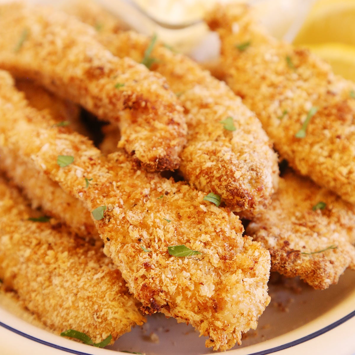 Chicken tenders on a platter sprinkled with fresh parsley flakes and a side of lemon wedges and sauce.