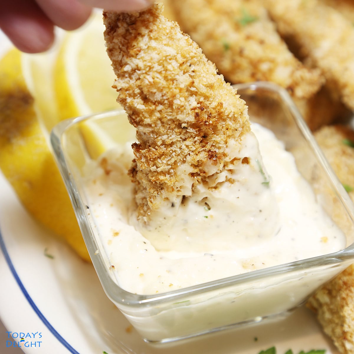 Chicken tender dipped in garlic aoili sauce and more tenders on platter with lemon wedges.