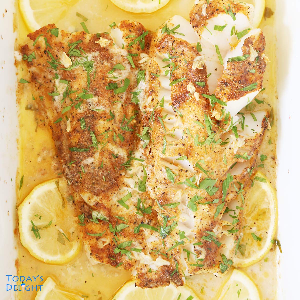 Baked cod fillet fish cut in half in garlic butter sauce garnished with parsley and surrounded with sliced lemon.