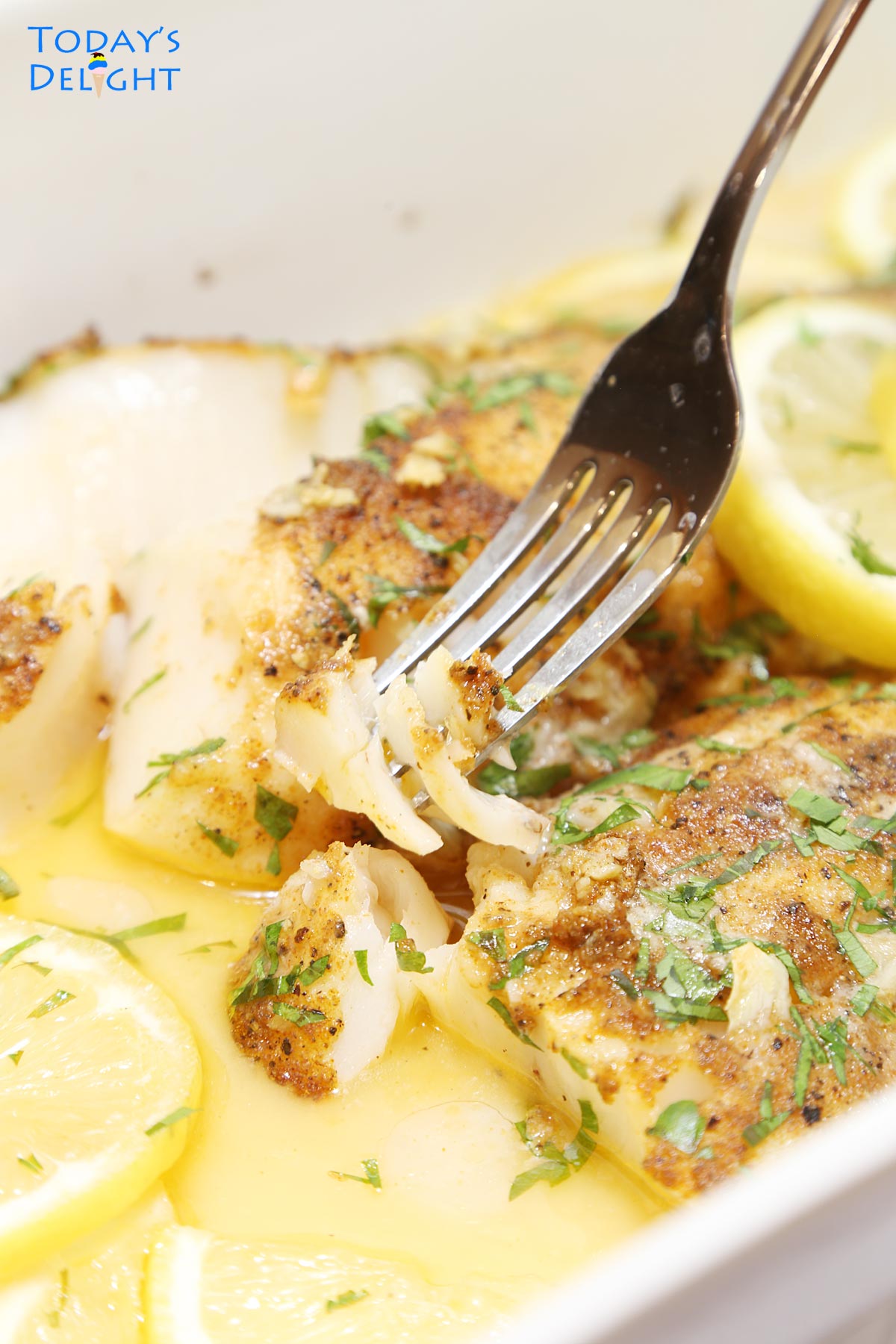Flaky baked cod in fork and casserole in garlic butter sauce garnished with parsley and lemon slices.