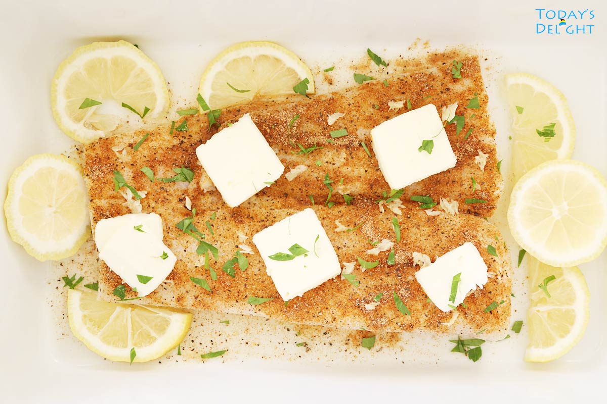 Cod fillet cut in half, seasoned and slices of butter on top and surrounded with sliced lemon in a casserole.