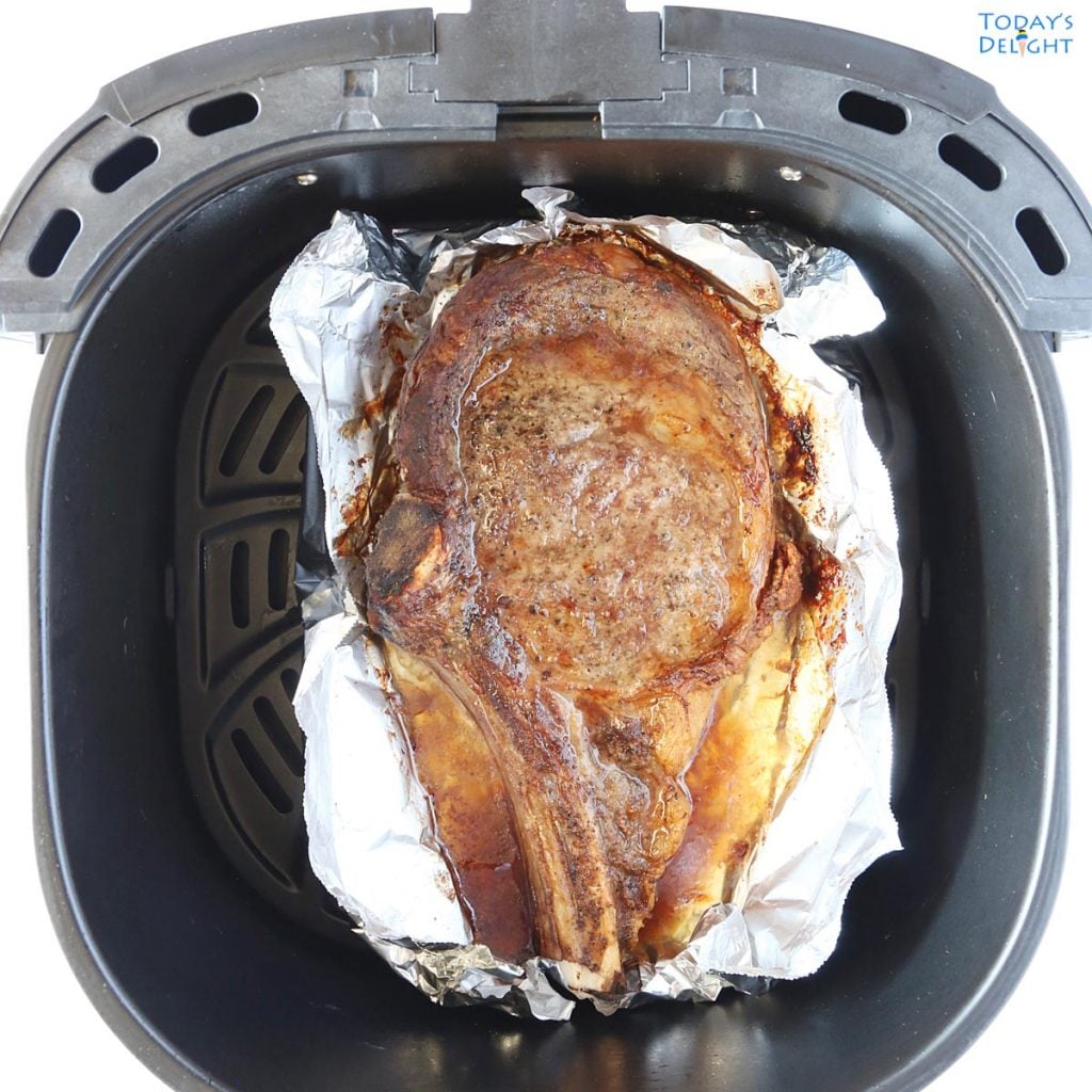 One cooked bone-in ribeye steak in air fryer basket lined with foil.