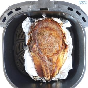 One cooked bone-in ribeye steak in air fryer basket lined with foil.
