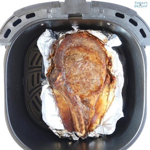 One cooked bone-in ribeye steak in air fryer basket lined with foil.