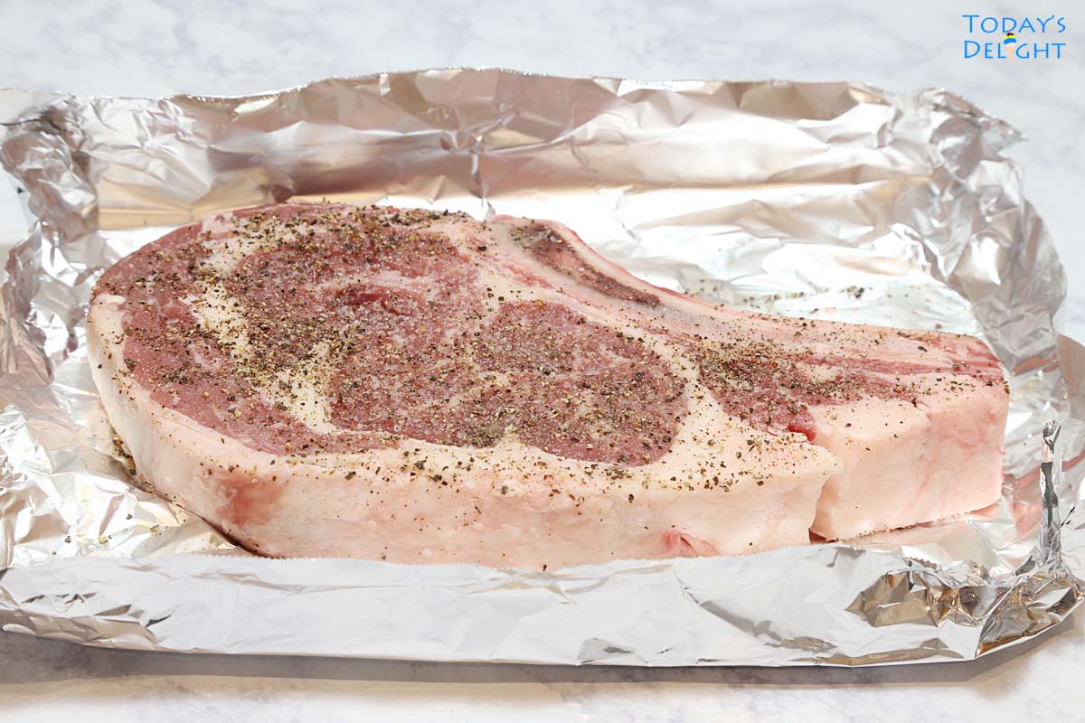 One uncooked bone-in ribeye steak seasoned with salt and pepper on a foil.