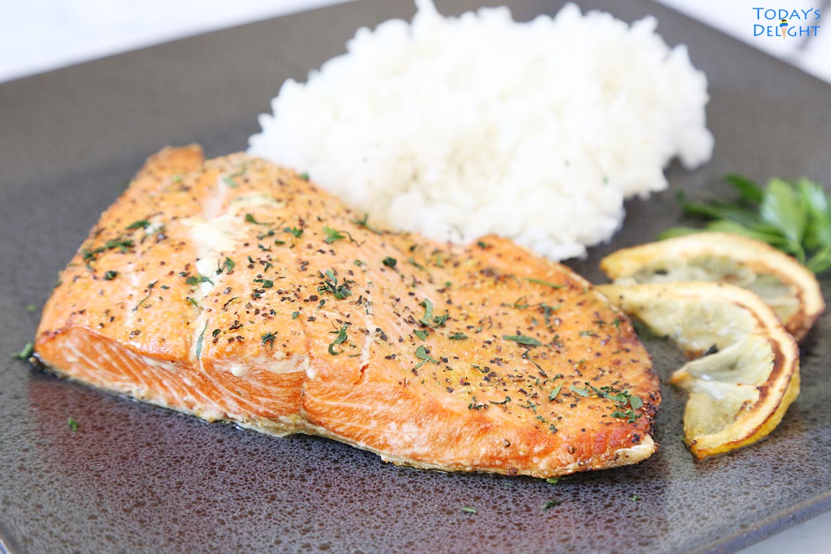 Air fried lemon pepper salmon with a side of white rice, 2 lemon wedges and some fresh parsley leaves.