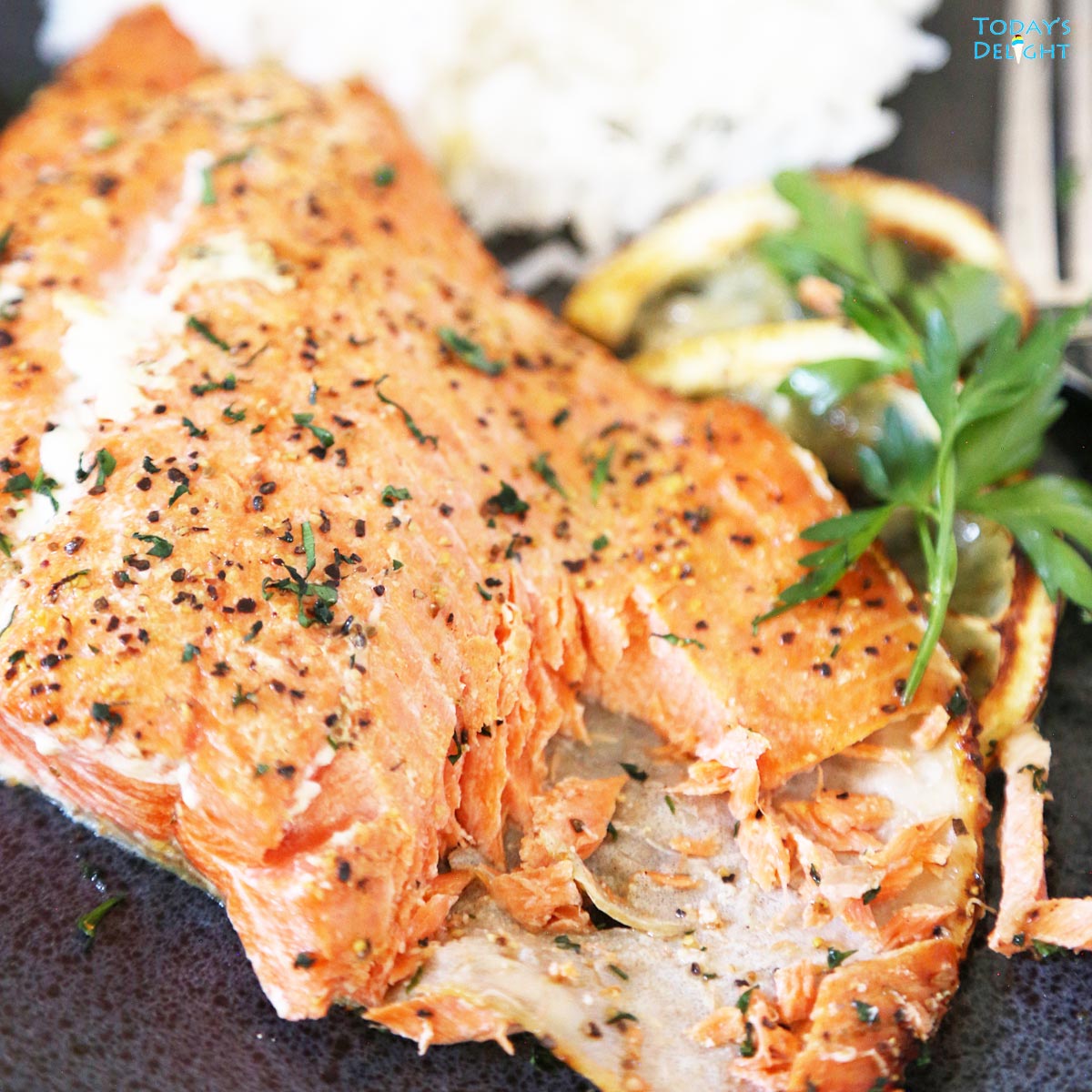 Part of cooked salmon flesh is removed to show the inside and a side of white rice, 2 lemons wedges and some fresh parsley leaves.