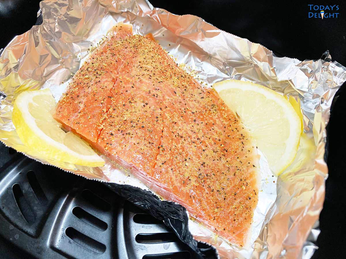 Raw seasoned salmon fillet with 2 lemon wedges on foil in air fryer basket.