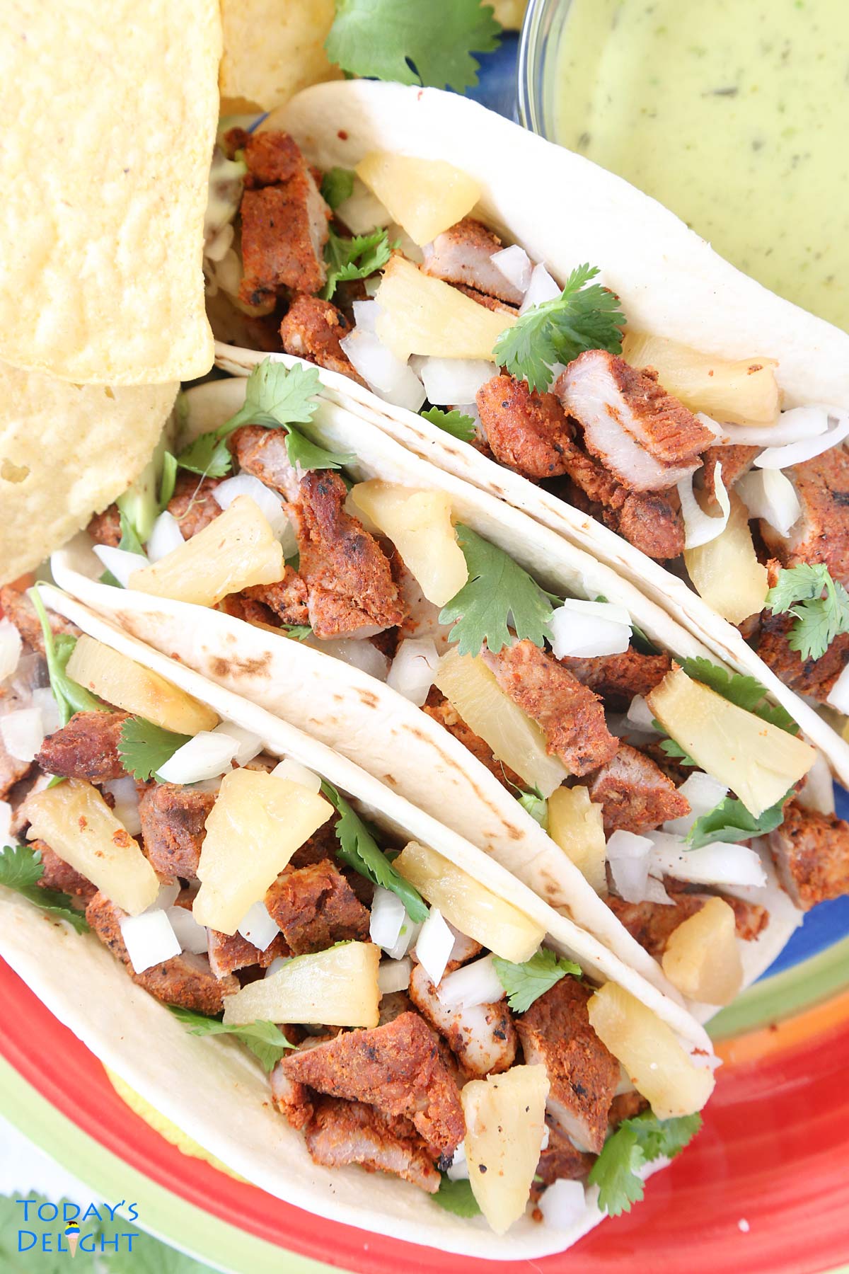 Three tacos al pastor on a plate with sides of tortilla chips, guacamole salsa and lime wedges.
