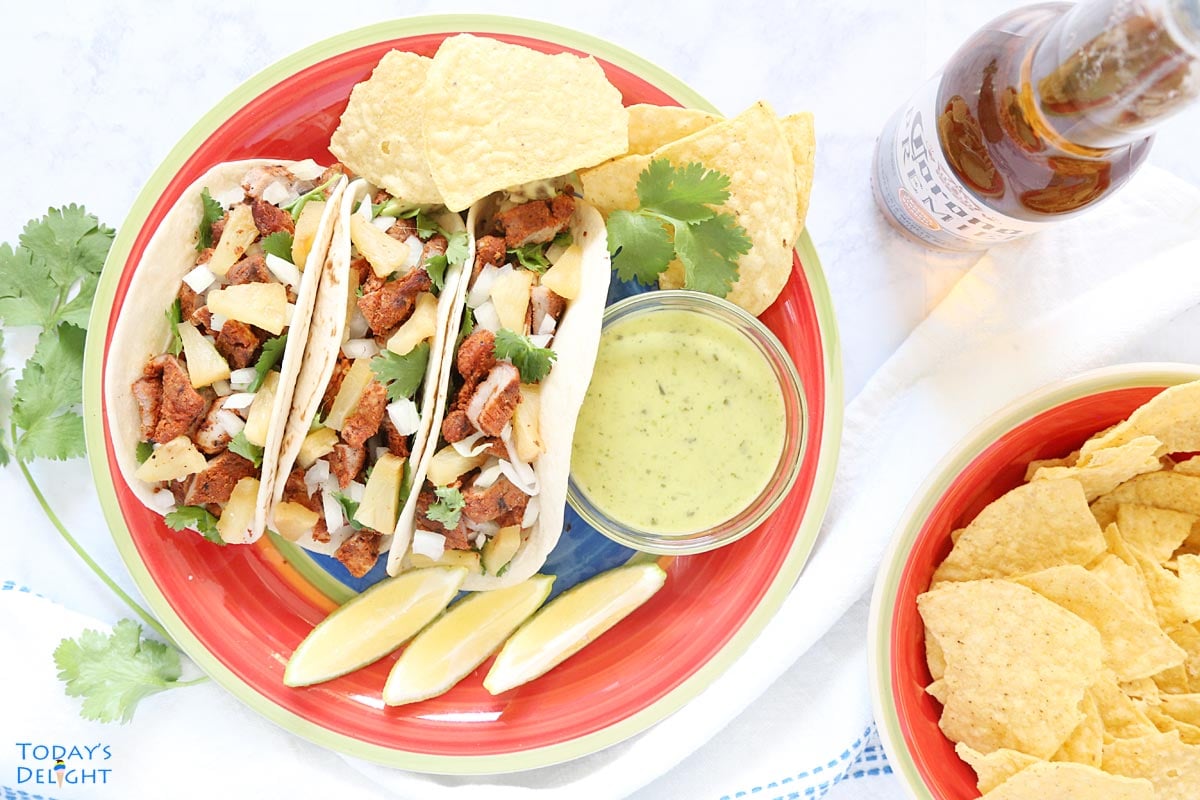 Three tacos al pastor on a plate with sides of nacho chips, guacamole salsa, lime wedges and beer.