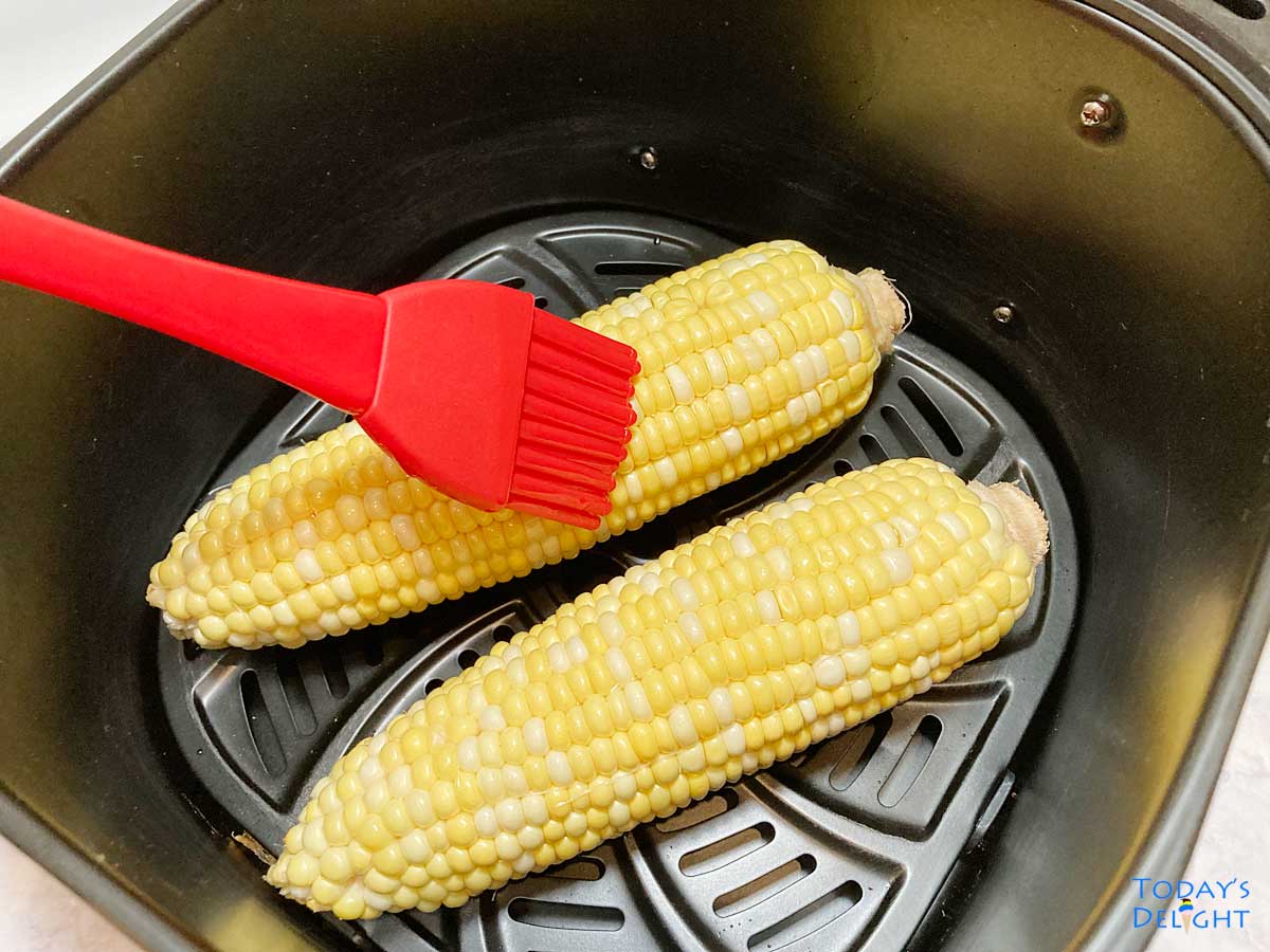 Two corn on the cob brushed with oil in air fryer basket.